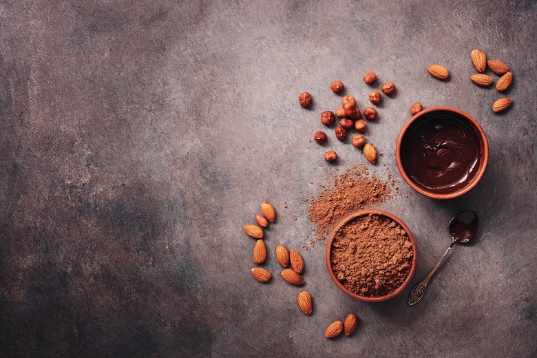 A bowl of melted chocolate, cocoa powder, almonds and hazelnuts on a dark rustic background.