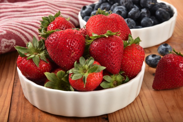 A bowl of fresh ripe strawberries with blueberries in the background.