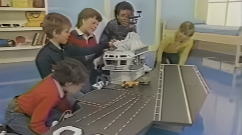Children gathered around a large toy aircraft carrier playset in a brightly lit playroom.