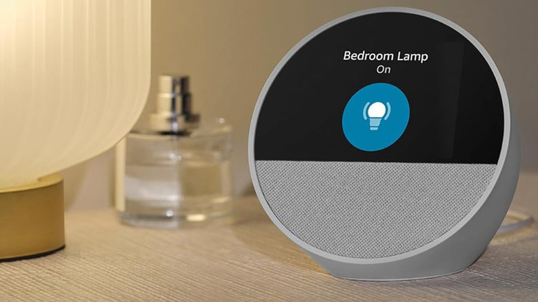 An Amazon Echo Show on a bedside table near a lamp.