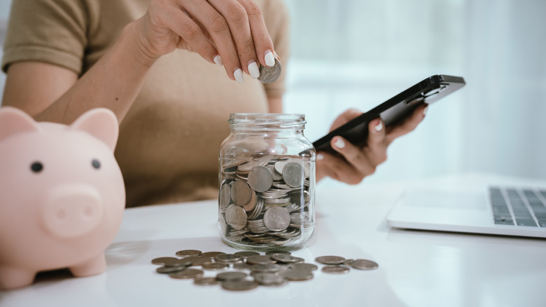 A person looking at phone and putting coins into a jar next to a piggy bank