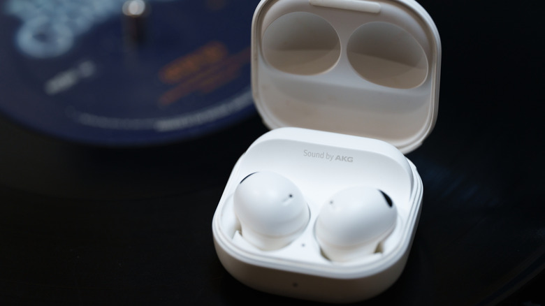 An open charging case containing a pair of white wireless earbuds, with Sound by AKG branding visible inside the case and a blurred audio device in the background.