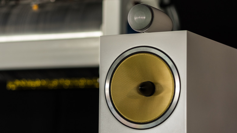 Close-up of a high-end bookshelf speaker featuring a yellow woven-cone woofer and a separate metal tweeter mounted on top, with a softly blurred audio setup in the background.