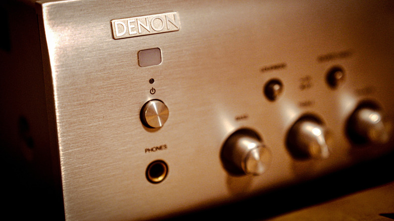 Close-up of a Denon stereo amplifier front panel, showing the power button, headphone jack, and control knobs on a brushed aluminum faceplate.