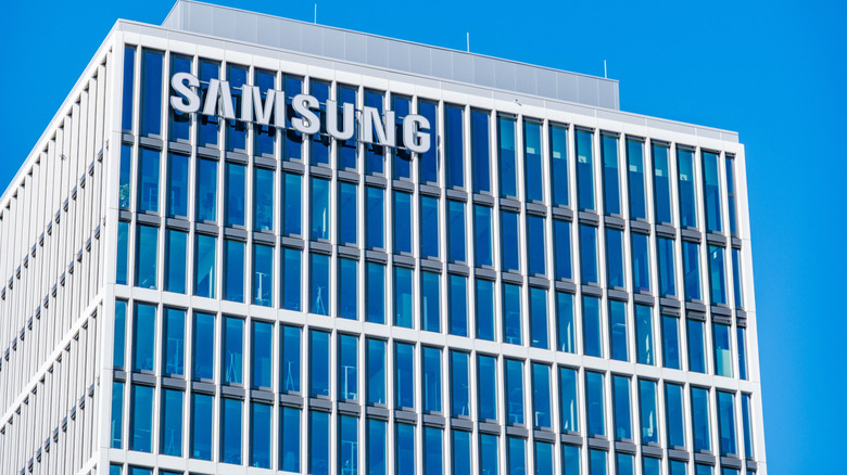A Samsung corporate office building with the company logo displayed prominently on the glass facade against a clear blue sky.