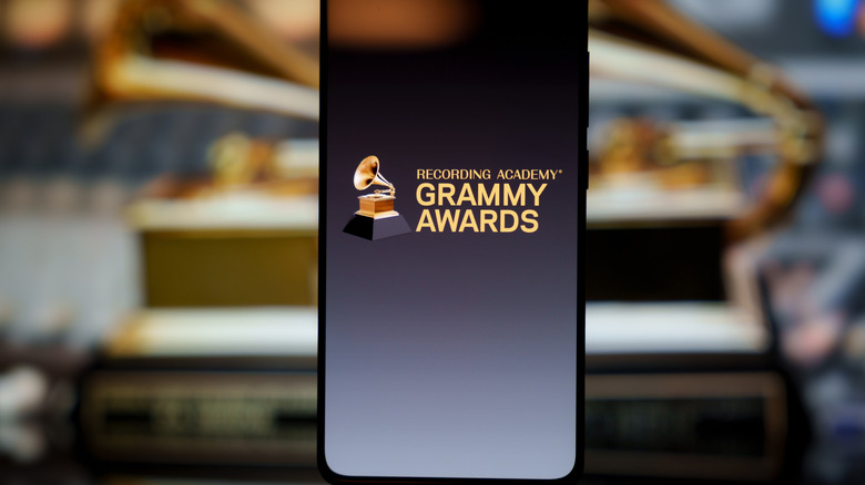 A smartphone displaying the Recording Academy Grammy Awards logo, with a blurred Grammy trophy visible in the background.