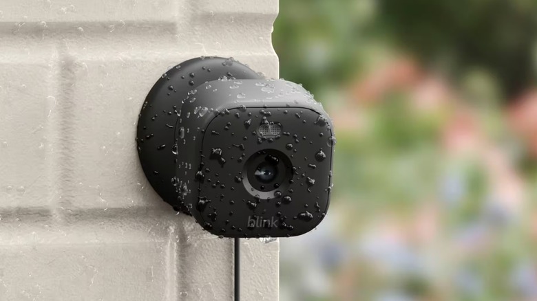 The Blink Mini 2 security camera installed outdoors on a white brick wall and wet with rain.