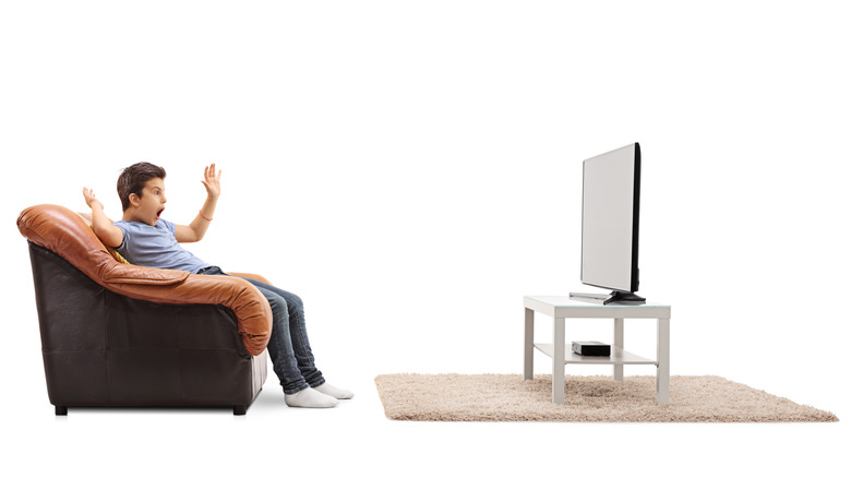 A young man sitting on a couch watching TV with his arms in the air, against a white background.
