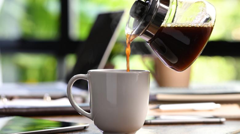 A person pouring coffee into a mug with a laptop in the background.