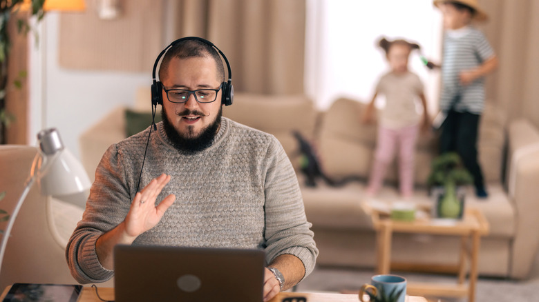 A person with a headset using a laptop while kids stand on a couch in the background.