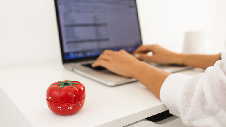 A person working on a laptop with a timer for the Pomodoro technique on a desk.