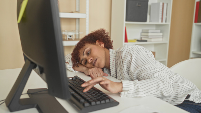 A woman resting her head on a desk while typing on a keyboard.