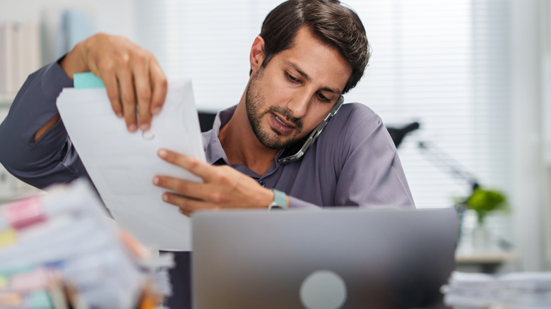 A man on a phone call while handling documents in front of a laptop.