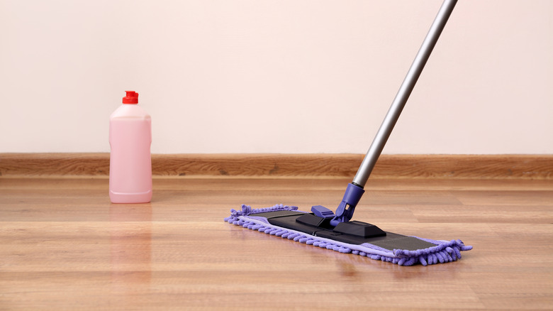 A bottle of pink cleaning liquid beside a stick mop with a purple microfiber pad on a light brown hardwood floor
