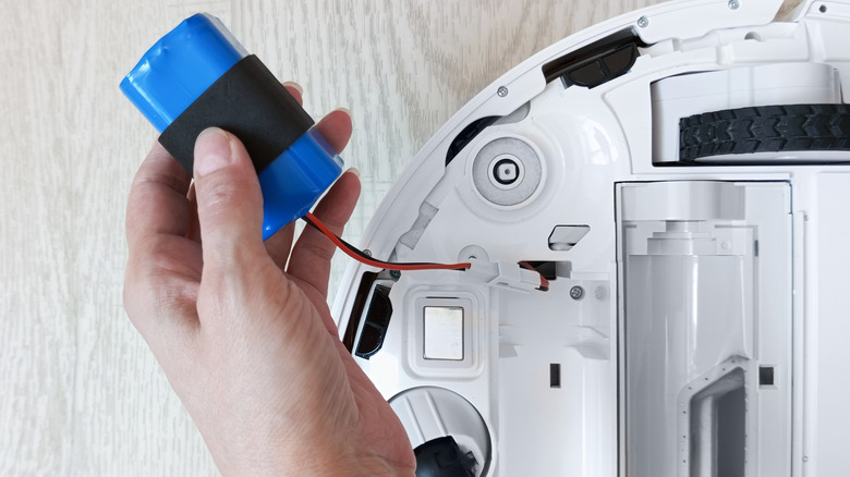 A hand holding a blue battery pack connected to the underside of a white robot vacuum against a light gray wooden floor