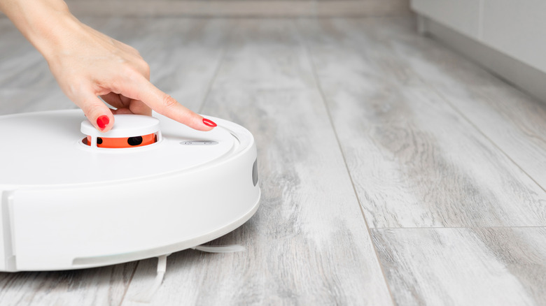 A manicured hand hovering a finger over a white robot vacuum placed on a light gray floor