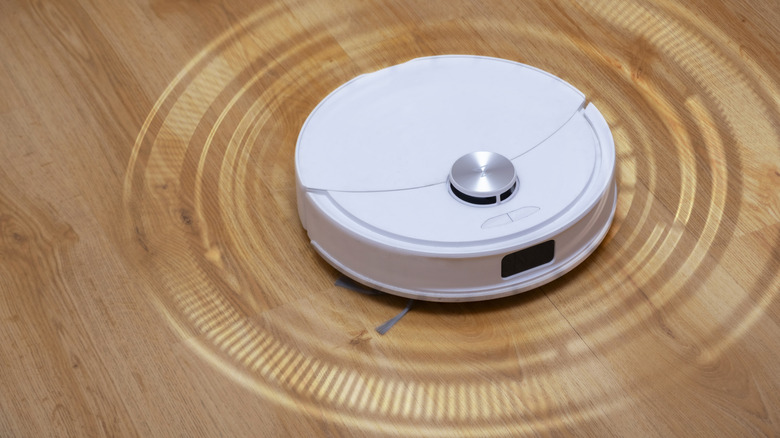 A white robot vacuum scanning its surroundings and projecting rings of light on a tan hardwood floor