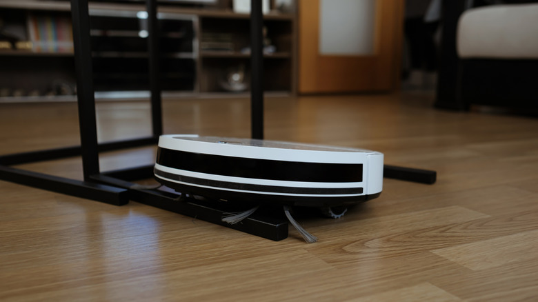 A black and white robot vacuum stuck on horizontal black table legs on a light brown hardwood floor
