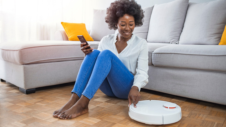 Woman on a brown hardwood floor holding a smartphone and pressing a button on a white robot vacuum