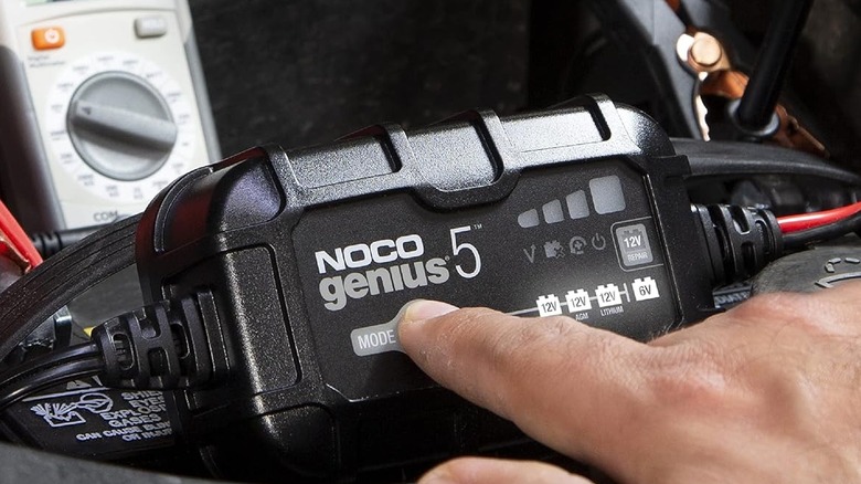 A hand using a Noco Genius 5 car battery charger in a car engine bay.