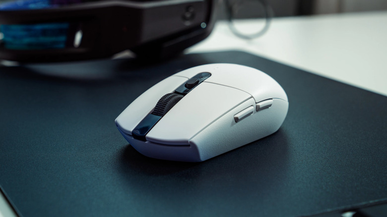 A white wireless gaming mouse on a black mouse pad, with blurred desktop equipment in the background.