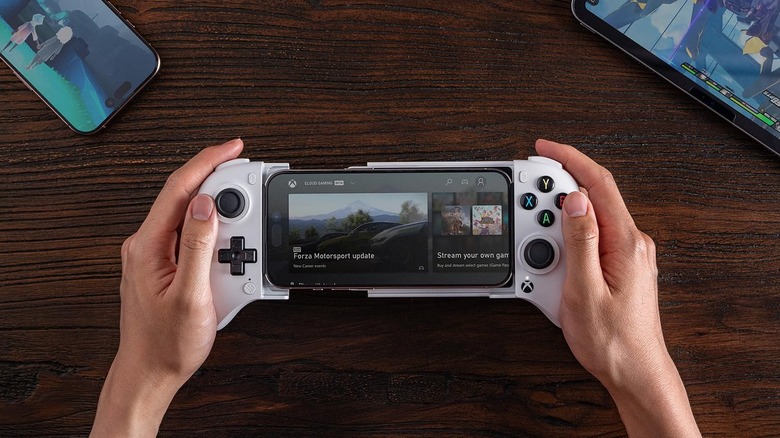 Hands holding a white mobile gaming controller with a smartphone attached in the center, displaying the Xbox Cloud Gaming interface on a wooden table.