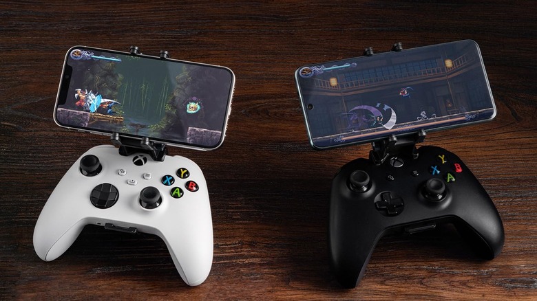 Two Xbox controllers, one white and one black, each fitted with a smartphone clip holding a phone displaying a side-scrolling action game on a wooden surface.