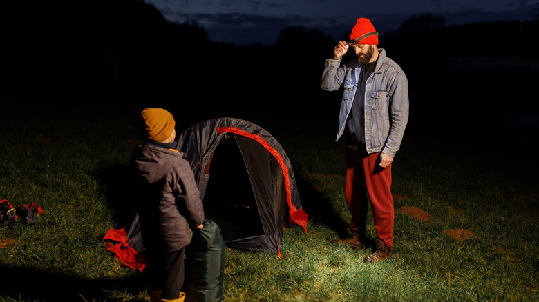 A father and his young child at a camping ground. The man is fiiddling with his headlamp.