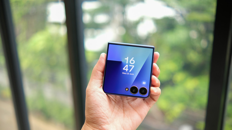 A folded Samsung Galaxy Z Flip7 smartphone, held against a window overlooking greenery