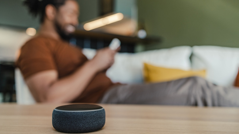 An Amazon Alexa speaker on a wooden table with a smiling person blurred in the background