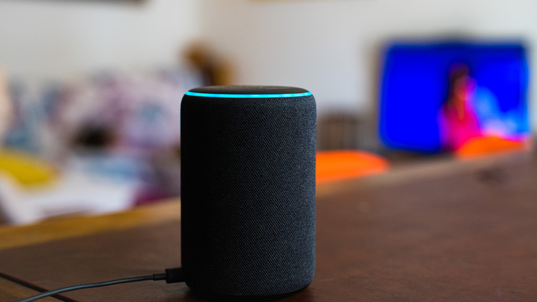 An Alexa speaker with a wire connected to it, placed on a wooden surface with blurred people and objects in the background