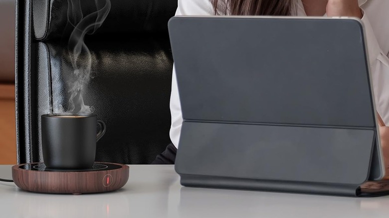 The Sweetfull MW-07 coffee mug warmer on a desk, beside a person working on a tablet.