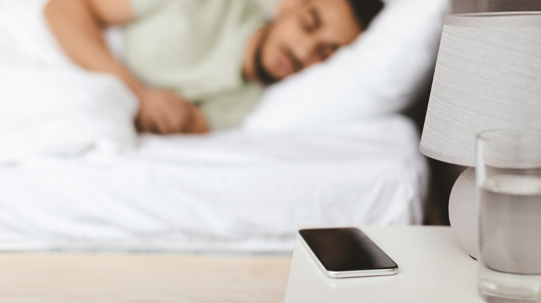 Close-up of a phone on a nightstand with a man lying in bed in the background