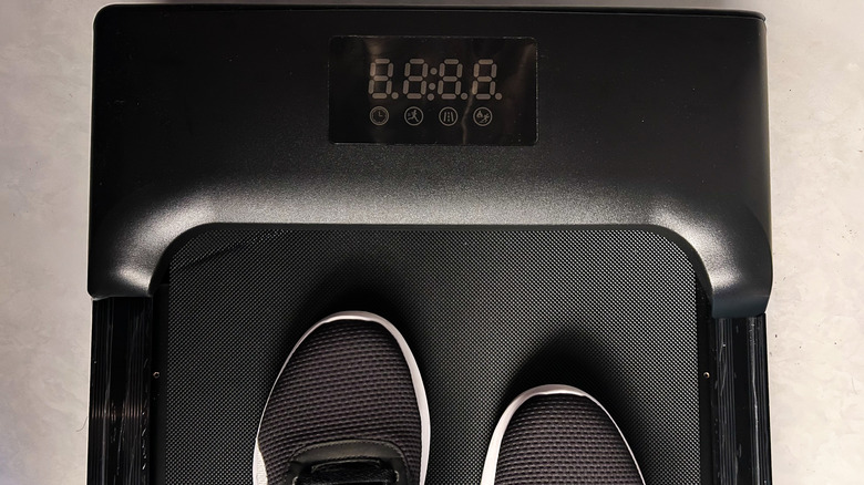 Top-down view of a walking pad treadmill with a digital display, showing a person standing on the belt in athletic shoes.