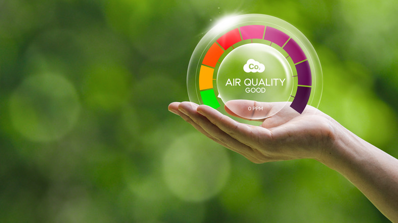 Hand holding a digital air quality gauge displaying a "Good" air quality rating against a blurred green outdoor background.