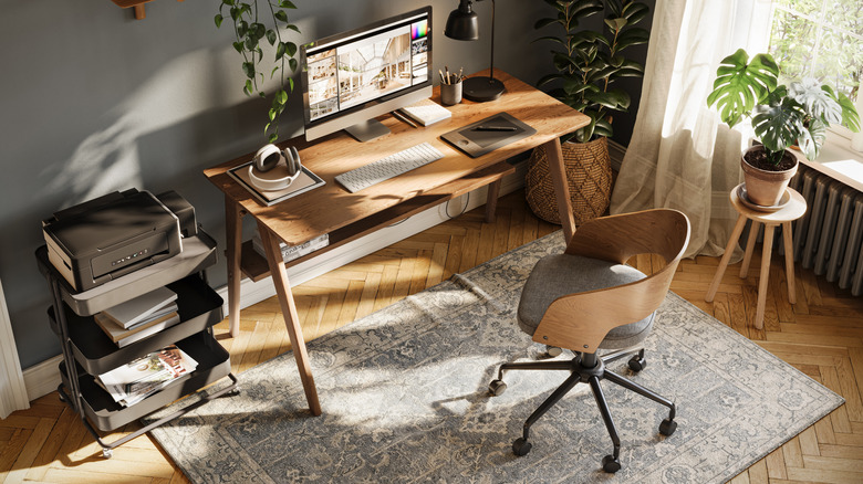 Bright and organized home office featuring a wooden desk with a desktop computer, drawing tablet, headphones, printer cart, ergonomic chair, and several indoor plants illuminated by natural sunlight.