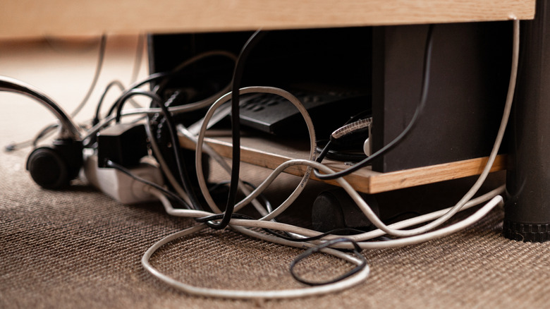 Close-up of tangled power and data cables beneath a desk, with a power strip and electronic devices creating cable clutter.