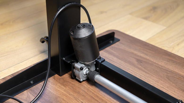 Close-up of the electric motor and lifting mechanism mounted beneath a height-adjustable standing desk, showing the drive system attached to the desk frame and support leg.
