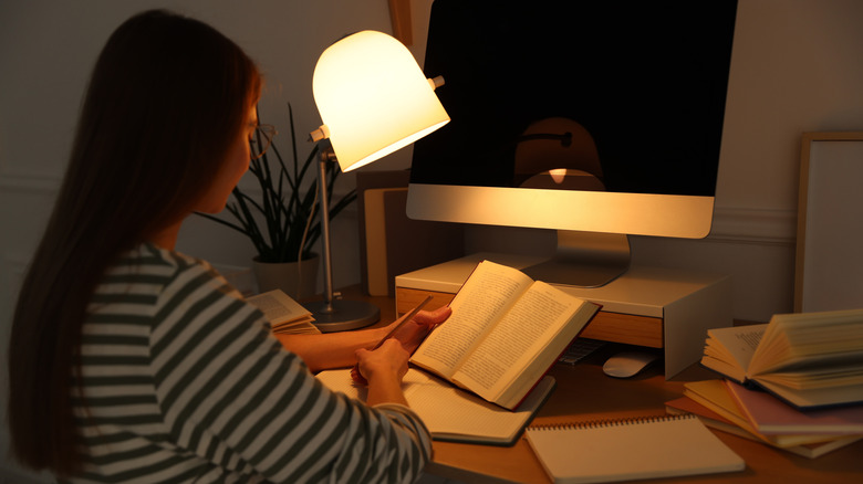 A woman sitting in a modern office using a desk lamp to illuminate her book and notes.
