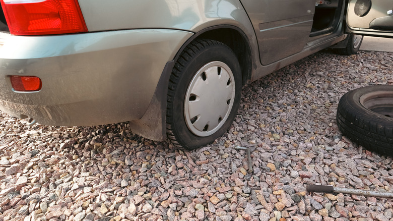 A car sits on a gravel driveaway with a deflated tire and replacement by the side