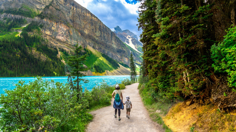 An adult and a child walking on a path in a park with water on one side and trees on the other side.
