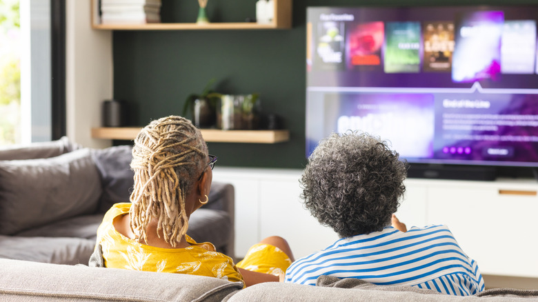 Two senior citizens sitting on the couch while operating a smart TV.