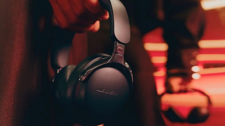 A pair of black Bose QuietComfort Ultra Headphones gen 2 being held by hand at person's side with dark background