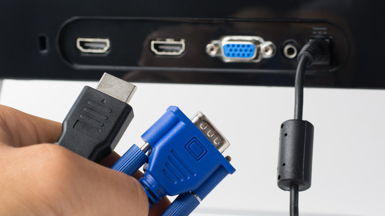 A hand holds a VGA and HDMI cable with the rear of a monitor in the background.