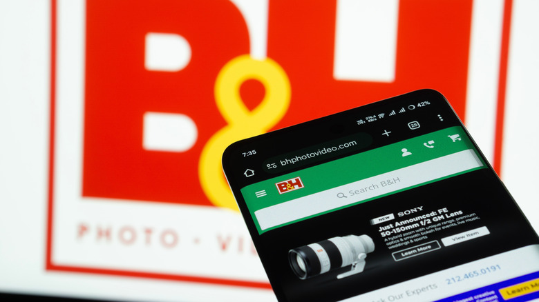 The B&H Photo Video website on a smartphone, with the retailer's logo on a screen in the background.