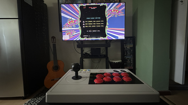 A TV displaying retro games on a rolling stand with an arcade stick in the foreground.