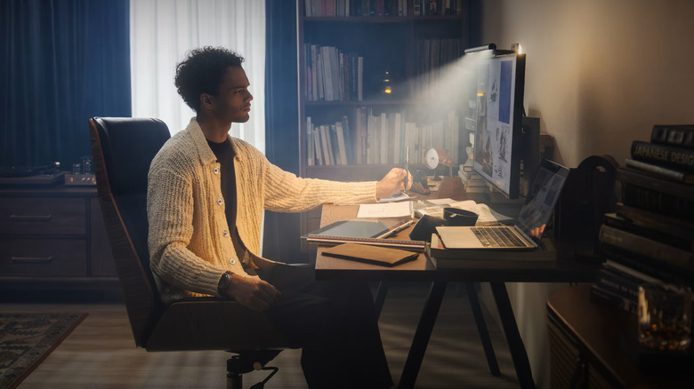 A man sitting at a desk with a laptop and monitor. There is a BenQ monitor lightbar attached the monitor which is lighting the workspace.