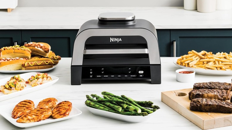 The Ninja DG551 Foodi Smart XL 6-in-1 indoor grill on a kitchen countertop, surrounded by different kinds of food.