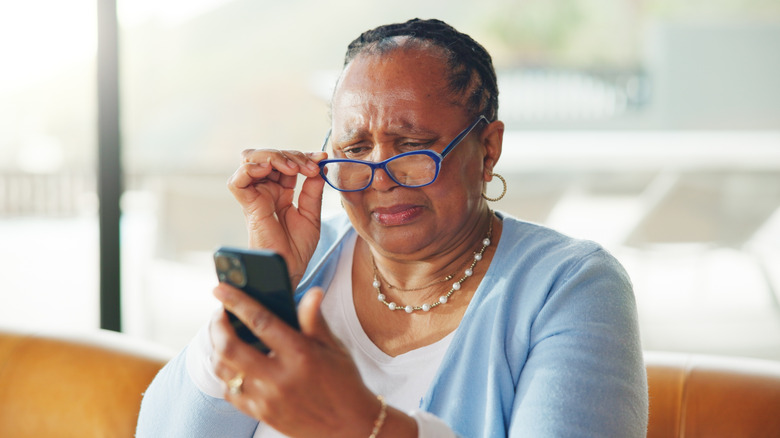 A person with a quizzical expression, adjusting their glasses as they look at a phone in their hand
