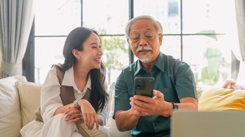 An old person using their phone as a younger person watches on with a smile